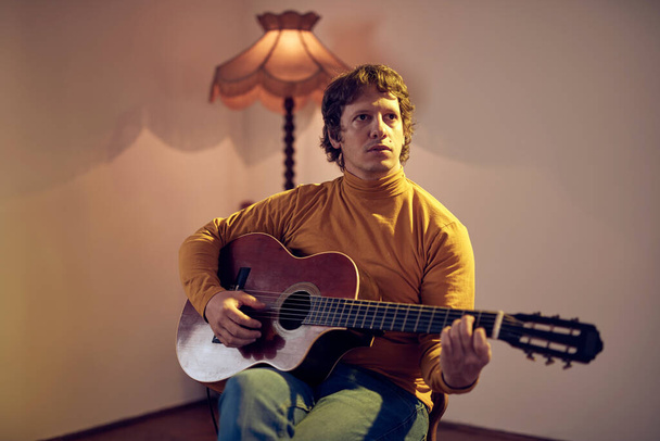 Mužský hudebník hrající na akustickou kytaru na zesilovači v retro vintage místnosti. - Fotografie, Obrázek