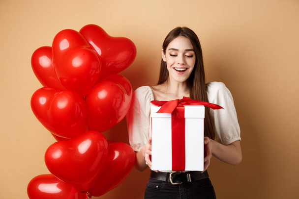 Happy woman open Valentines day gift, usměvavý vzrušený a při pohledu na současnost box, stojící v blízkosti červené srdce balónky na béžové pozadí - Fotografie, Obrázek
