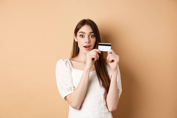 Plastik kredi kartı gösteren heyecanlı genç bir kadın bej arka planda duran kameraya hayret ediyor. - Fotoğraf, Görsel