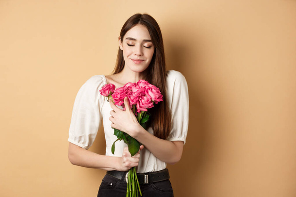 Valentines day. Tender and romantic girl, smell roses and smile with closed eyes. Girlfriend hugging gift flowers from lover, standing on beige background - Photo, Image