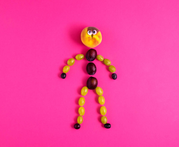 Een menselijke figuur gemaakt van druivenkorrels, pruimen en perzik met ogen op pastel heldere paarse achtergrond. Minimalistisch creatief herfstconcept, vlakke lay compositie - Foto, afbeelding
