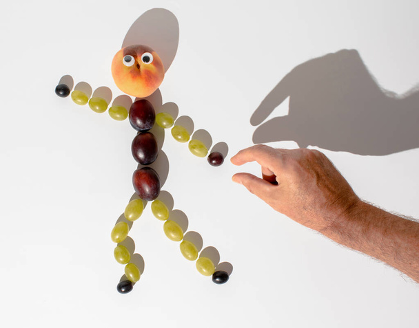 Людська фігура з виноградних зерен, слив і персиків тікає від чоловічої руки. Мінімалістична креативна кумедна концепція осені, плоска композиція на білому тлі
 - Фото, зображення