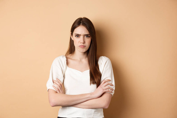 Angry girlfriend cross arms on chest and frowning, looking offended or disappointed, sulking at you, standing mad on beige background - Photo, Image