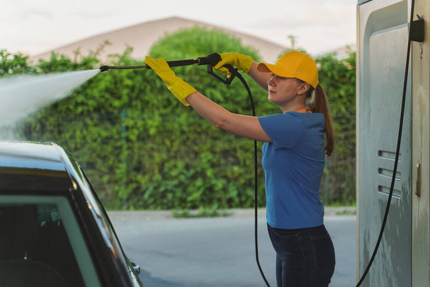 Woman in uniform cleaning car using high pressure water. Car wash service. - Photo, Image