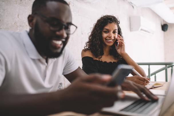Portrait of dark skinned female freelancer smiling at camera during collaboration meeting with successful man, happy woman using mobile technology for making cellphone conversation via application - Photo, Image