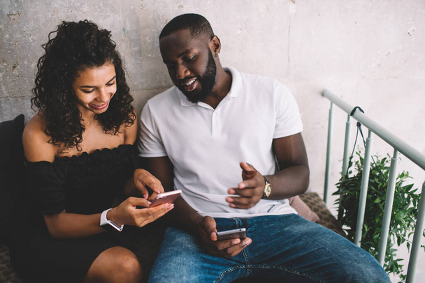Cheerful male and female friends using cellphone device for online messaging discussing funny social publication during cellular networking, happy dark skinned bloggers browsing website on leisure - 写真・画像