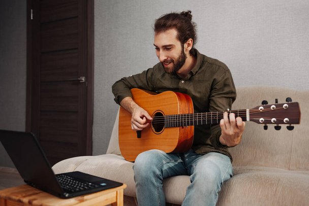 Young smiling man learning to play guitar at home and looking into laptop - Фото, изображение
