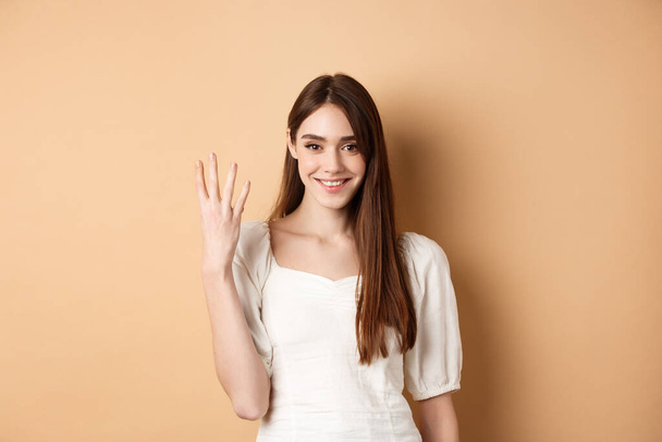 Приваблива молода жінка показує пальці номер чотири, посміхаючись і виглядаючи впевнено, стоїть на бежевому фоні
 - Фото, зображення