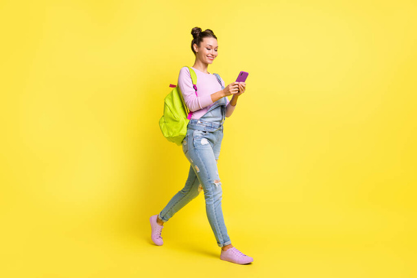 Photo of funny pretty young lady dressed jeans overall backpack typing modern gadget walking isolated yellow color background - Foto, Bild