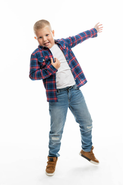 A cheerful boy 6-7 years old imitates playing the guitar. Schoolboy in jeans, a plaid shirt and a white T-shirt. White background. Vertical. - Photo, Image