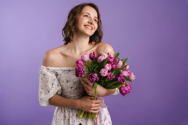 White young woman smiling while posing with tulips isolated over purple wall - 写真・画像