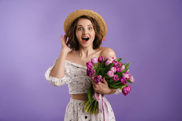 White surprised woman in hat exclaiming while posing with tulips isolated over purple wall - Фото, изображение