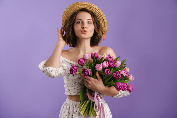 White young woman in hat making kiss lips while posing with tulips isolated over purple wall - Foto, Bild