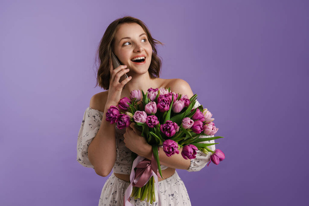 Smiling woman talking on cellphone while posing with tulips isolated over purple wall - Photo, Image