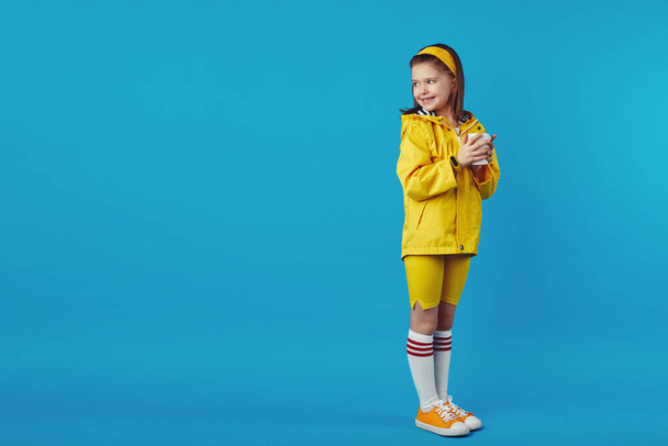 Little cute girl wears raincoat holds takeaway cup with tea and looking aside - Foto, Imagem
