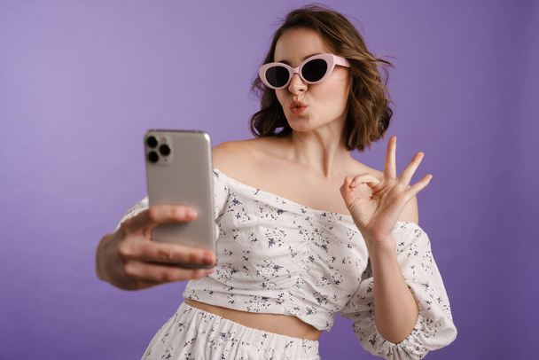 Feliz joven mujer blanca sosteniendo el teléfono móvil de pie sobre fondo violeta, tomando una selfie, usando gafas de sol - Foto, Imagen