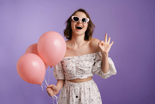 Excited young woman gesturing while posing with balloons isolated over purple wall - Foto, immagini