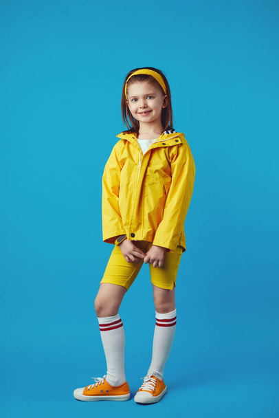 Kleines Mädchen in gelbem Regenmantel und Turnschuhen lächelt und hält die Hände zusammen - Foto, Bild