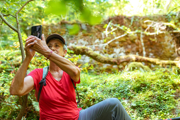 mujer mayor con pelo gris, gorra y camiseta roja tomando una selfie con su teléfono en el bosque - Foto, Imagen