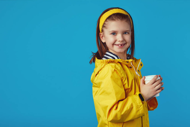 Mädchen trägt Regenmantel, hält Imbissbecher mit Tee und lächelt gegen Blau - Foto, Bild