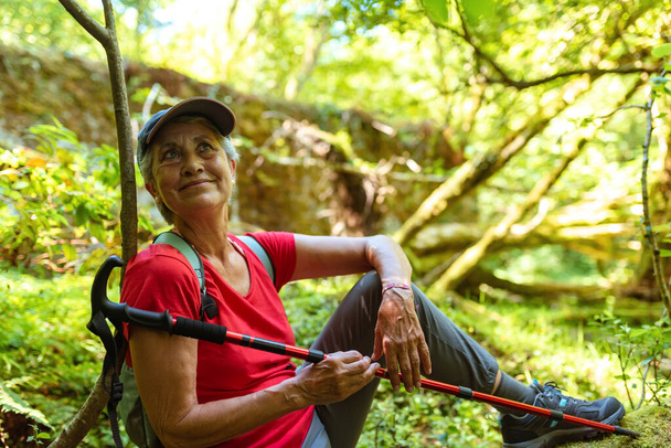 oudere vrouw met pet en rood t-shirt neemt een pauze tijdens een wandeling in het bos - Foto, afbeelding