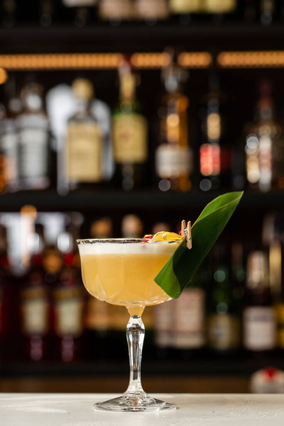 Glass with yellow lemon cocktail decorated with dried pineapple and mint on the blurred bar counter background. - 写真・画像