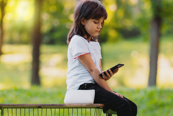 Фотографія красивої молодої дівчини, що сидить на лавці, тримає телефон у руці і переглядає цікаву інформацію
. - Фото, зображення