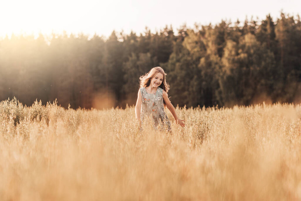 Ένα χαρούμενο κοριτσάκι τρέχει μέσα από ένα χωράφι με σιτάρι το καλοκαίρι μια ηλιόλουστη μέρα. Καλοκαίρι. Καλοκαιρινές διακοπές. Ευτυχισμένη παιδική ηλικία. Θετικά συναισθήματα και ενέργεια - Φωτογραφία, εικόνα