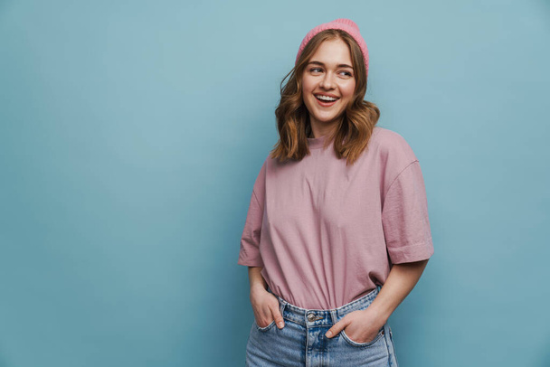 Young happy woman posing at camera with hands in pockets isolated over blue background - Foto, Bild