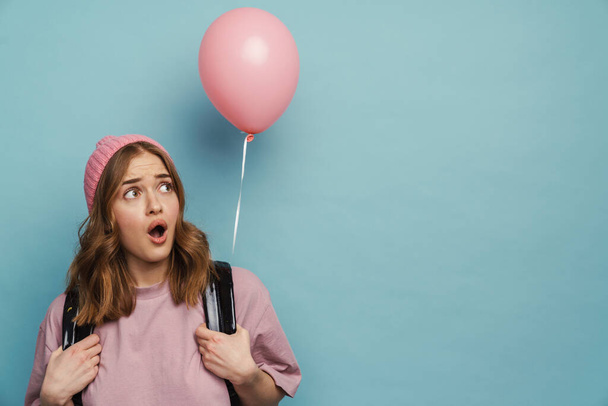 Young scared student girl expressing surprise while posing with balloon isolated over blue background - Фото, зображення