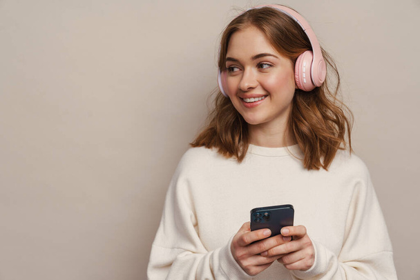 Young smiling woman listening music with headphones and cellphone isolated over grey background - Φωτογραφία, εικόνα