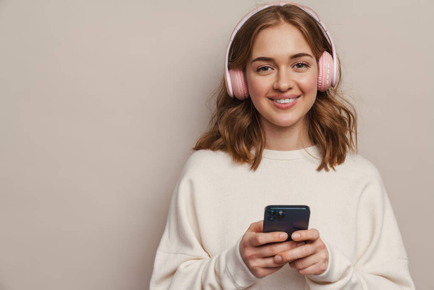 Young smiling woman listening music with headphones and cellphone isolated over grey background - Photo, image