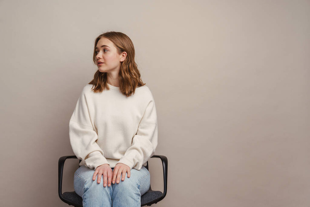 Young white woman looking aside while sitting on chair isolated over grey background - Foto, Imagem