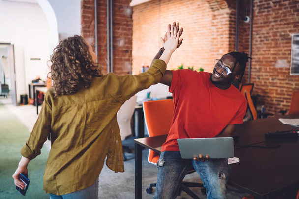 Happy youthful diverse coworkers in casual clothes rejoicing success of business project and giving high five during work together in loft open office - Foto, afbeelding