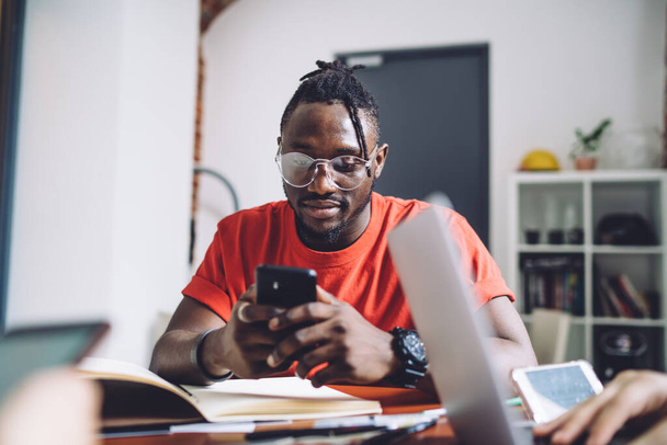 Serious African American male in casual red shirt and eyeglasses reading message on mobile phone while sitting at table with crop colleague - Foto, imagen