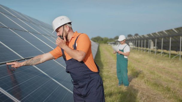 An employee of a power plant transmits commands by walkie-talkie. Happy caucasian adult engineer in helmet smiling at camera at solar power station outside. Ecological industry. Business team - Photo, Image