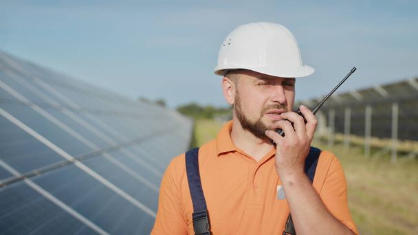 Щасливий кавказький дорослий інженер в шоломі посміхається на камеру на сонячній електростанції зовні. Екологічна промисловість. Концепція сонячної ферми. Співробітник електростанції передає команди по walkie-talkie
 - Фото, зображення