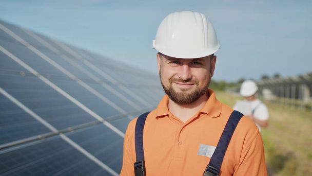 Счастливый белый взрослый инженер в шлеме улыбается в камеру на солнечной электростанции снаружи. Экологическая промышленность. Концепция солнечной фермы. Сотрудник электростанции передаёт команды по рации. - Фото, изображение
