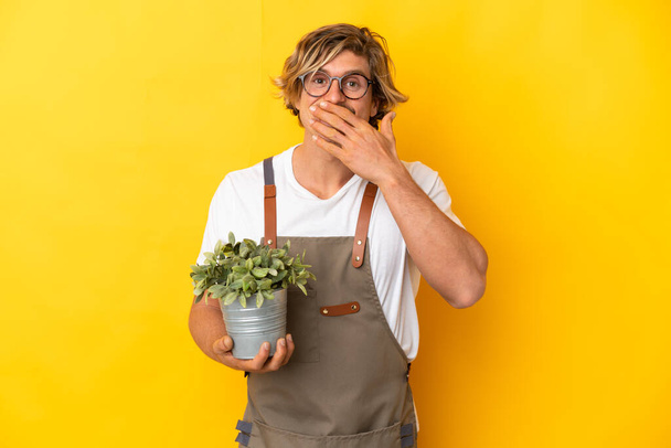 Gardener ξανθιά άνθρωπος κρατώντας ένα φυτό απομονωμένο σε κίτρινο φόντο χαρούμενος και χαμογελαστός κάλυψη στόμα με το χέρι - Φωτογραφία, εικόνα