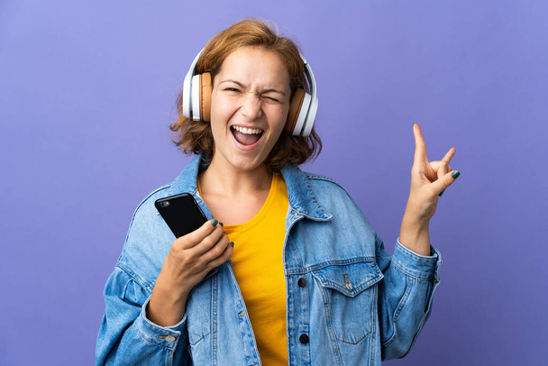 Fiatal grúz nő elszigetelt lila háttér hallgat zenét egy mobil, hogy rock gesztus - Fotó, kép