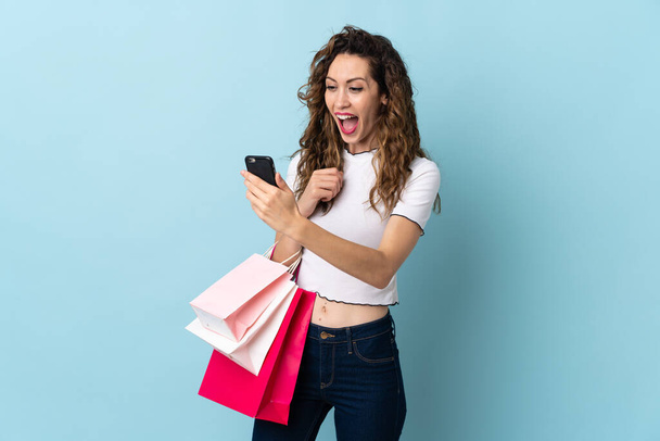 Jovem caucasiana isolada no fundo azul segurando sacos de compras e escrevendo uma mensagem com seu telefone celular para um amigo - Foto, Imagem