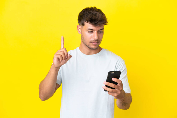 Joven caucásico guapo hombre aislado sobre fondo amarillo usando el teléfono móvil y levantando el dedo - Foto, imagen