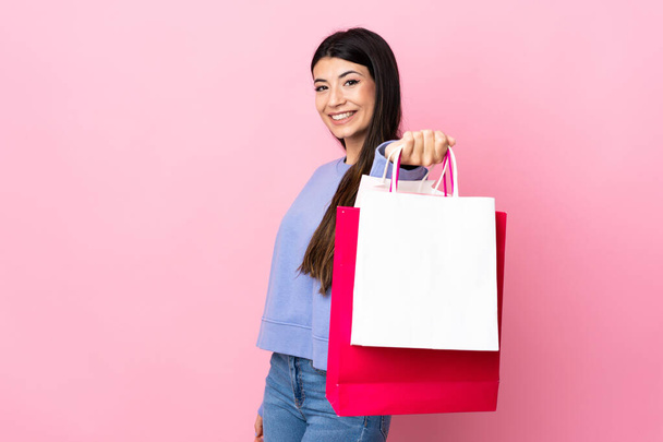 Молодая брюнетка на изолированном розовом фоне держит сумки с покупками и дает их кому-то - Фото, изображение