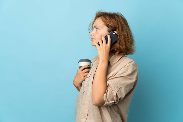 Young Georgian woman isolated on blue background holding coffee to take away and a mobile - Fotó, kép