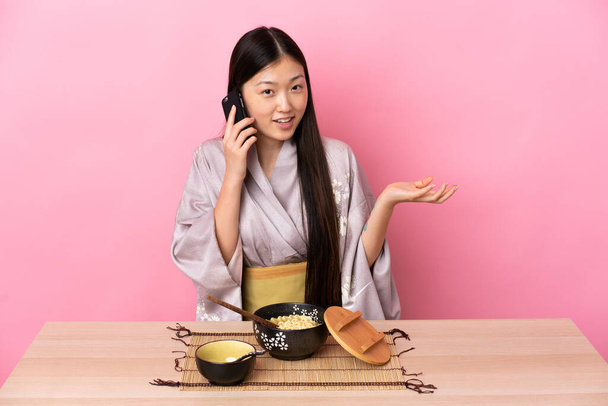 Junge Chinesin trägt Kimono und isst Nudeln, während sie mit jemandem auf dem Handy spricht - Foto, Bild