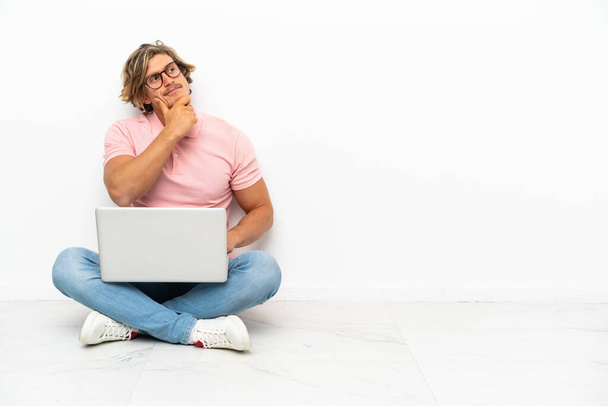 Young caucasian man sitting on the floor with his laptop isolated on white background having doubts - Foto, afbeelding