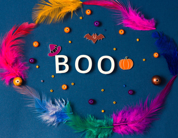 Boo word, colorful feathers on dark blue background. Halloween holiday concept. Bat, orange jack o lantern pumpkin, witch hat symbols. Party greeting card. Copy space, place for text. Flatly top view. - Photo, Image