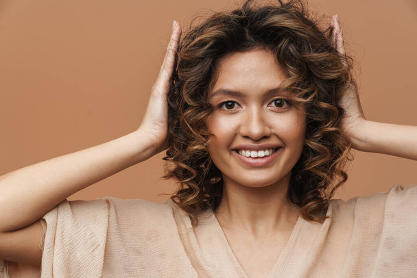 Young hispanic woman smiling while holding her head isolated over beige background - Фото, изображение