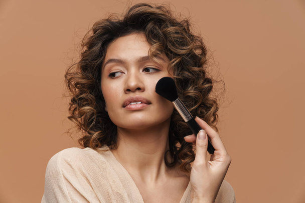 Young curly hispanic woman posing while using powder brush isolated over beige background - Photo, Image