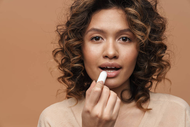 Young hispanic curly woman applying lipstick and looking at camera isolated over beige background - Foto, Bild